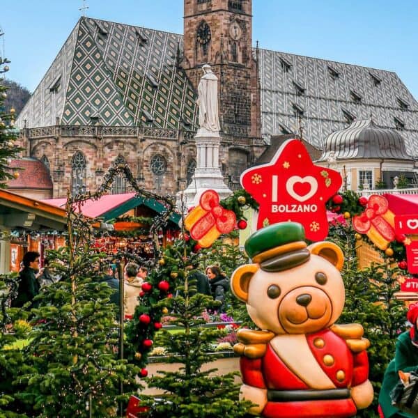 The festive Christmas market in Bolzano features charming decorated stalls, a grand teddy bear statue, and the historic cathedral beautifully framing the scene.