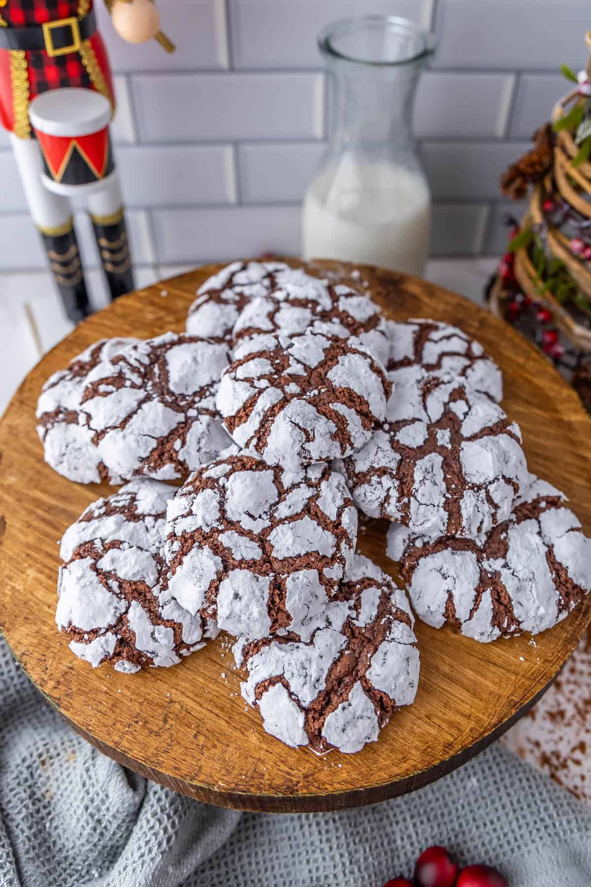 Chocolate crinkle cookies on a wooden stand with a bottle of milk and holiday decorations in the background.