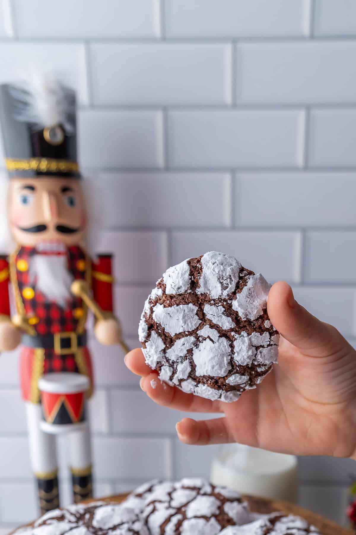 A hand holds a chocolate crinkle cookie dusted with powdered sugar. A nutcracker figurine is visible in the background against a tiled wall.