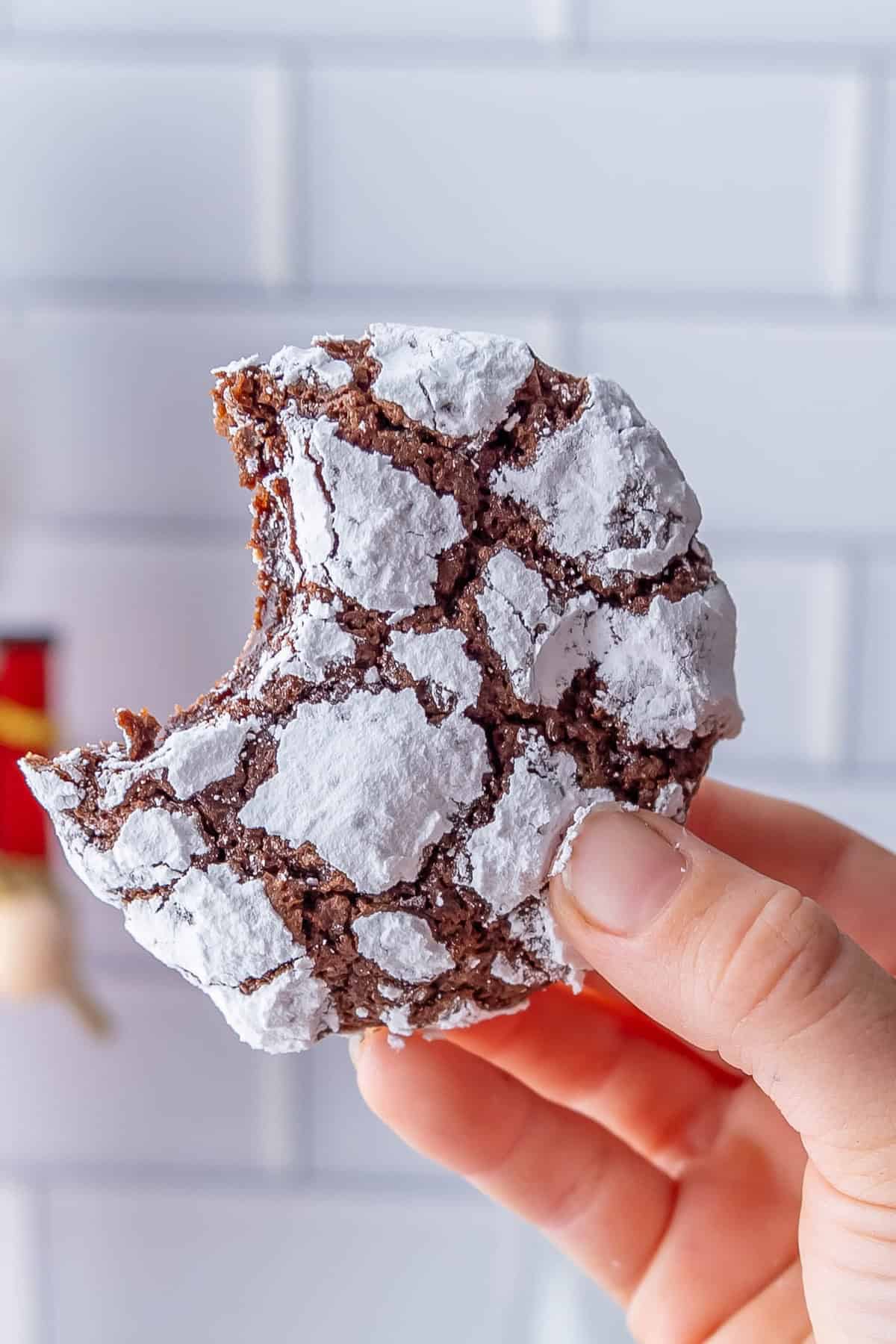 A hand holds a chocolate crinkle cookie with a bite taken out of it. The classic cookie, coated in powdered sugar, boasts its signature cracked appearance.