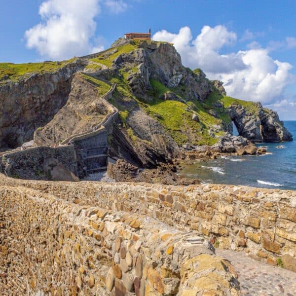 A stone path winds its way up to a small building atop a steep rock formation overlooking the sea under a partly cloudy sky—a must-see when you visit Basque Country.