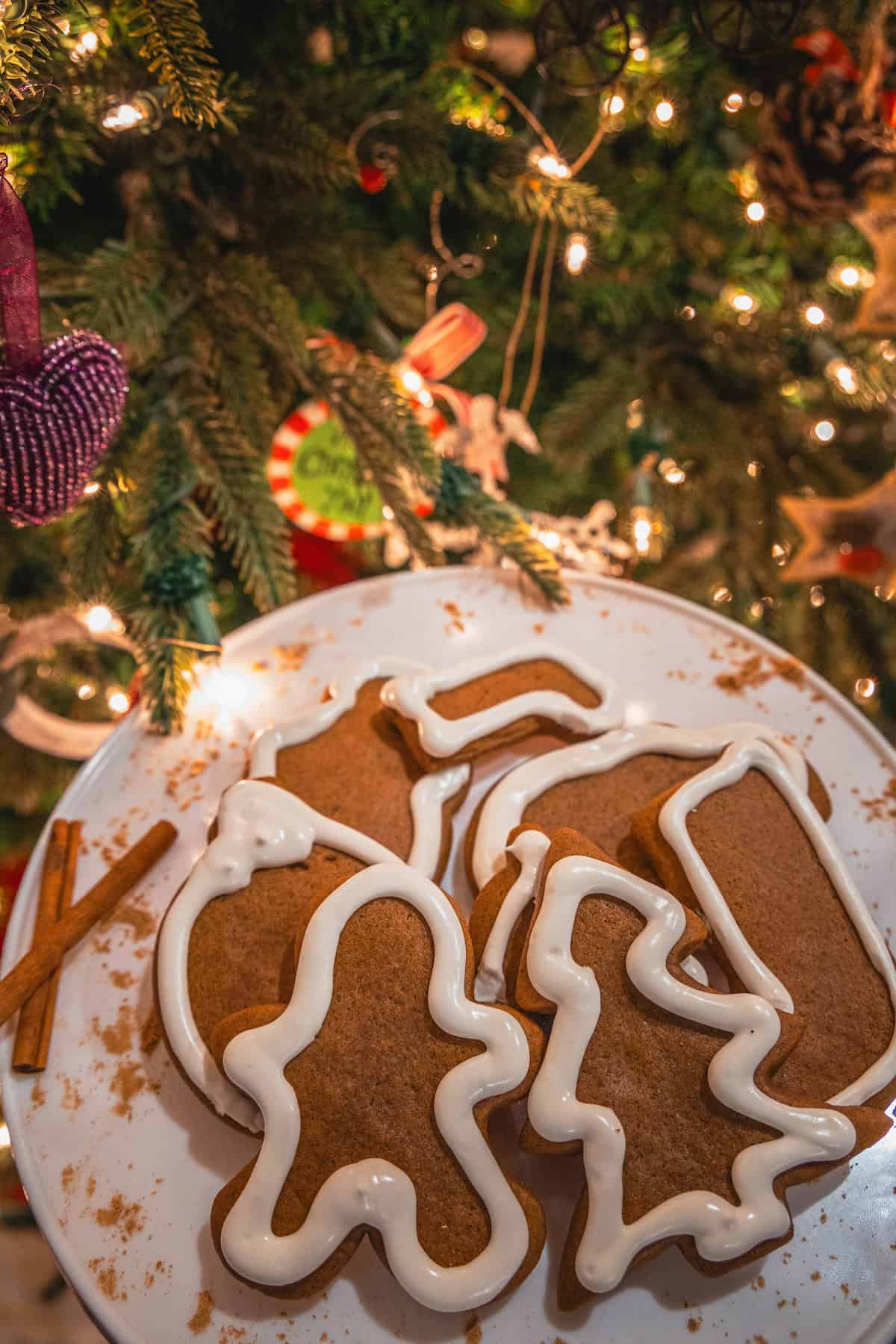 A charming plate of gingerbread cookies, intricately iced and shaped, sits elegantly near a resplendent Christmas tree adorned with twinkling lights and festive ornaments.