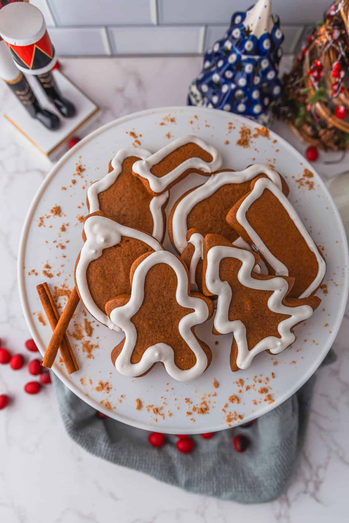 A plate of gingerbread cookies delicately shaped like a tree, gingerbread person, stocking, mitten, and ornament sits beautifully on a marble surface, with cinnamon sticks and cranberries artfully scattered around.