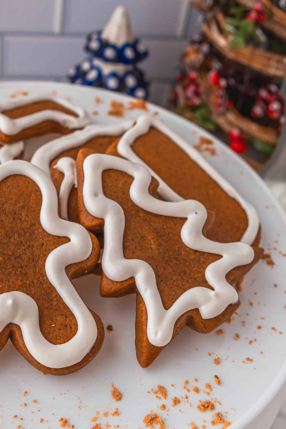 Gingerbread cookies with white icing in tree, candy cane, and gingerbread person shapes rest on a white plate, surrounded by crumbled gingerbread cookie bits.
