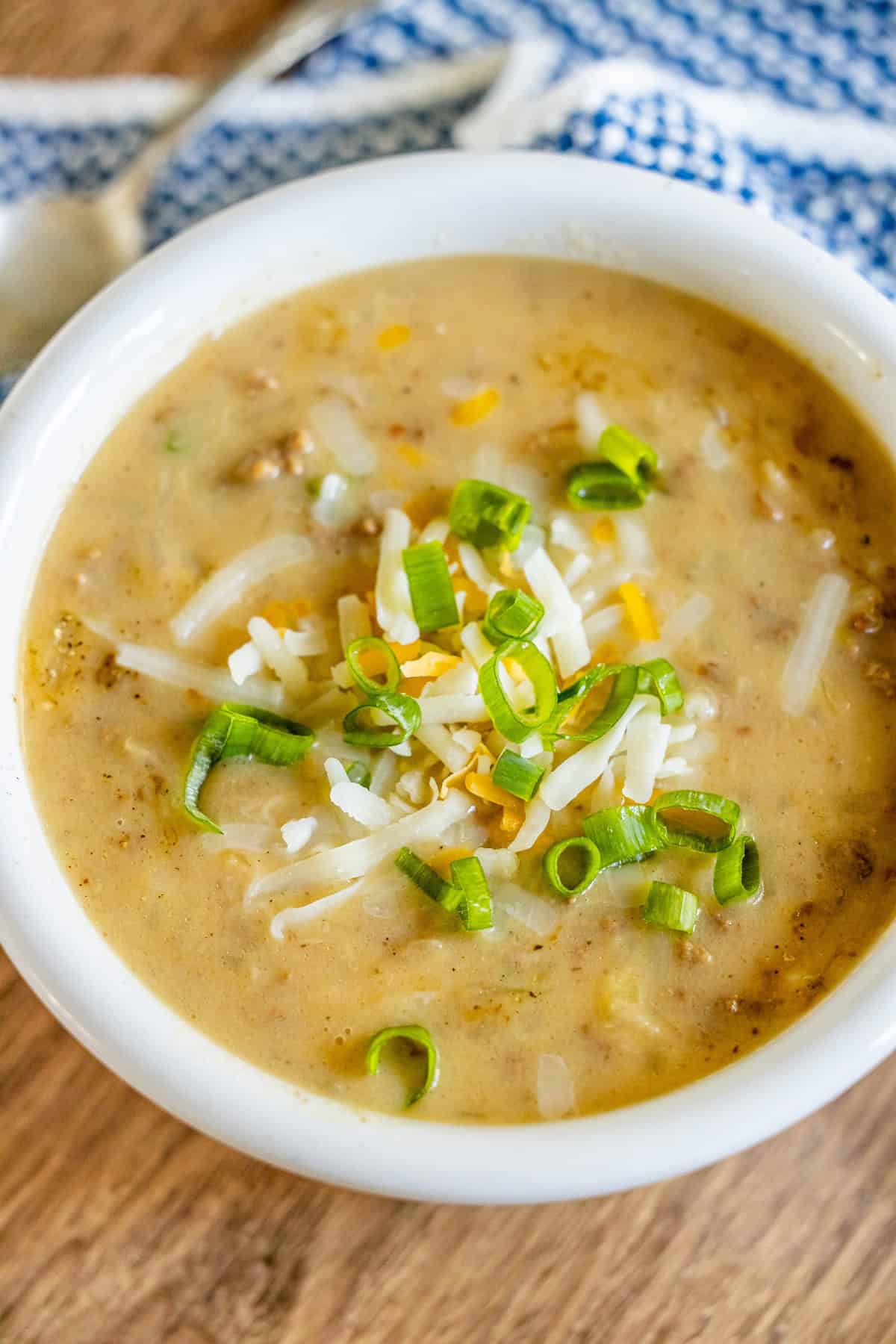 A bowl of creamy hash brown hamburger soup topped with shredded cheese and chopped green onions rests on a wooden surface, accompanied by a blue and white cloth and a spoon nearby.