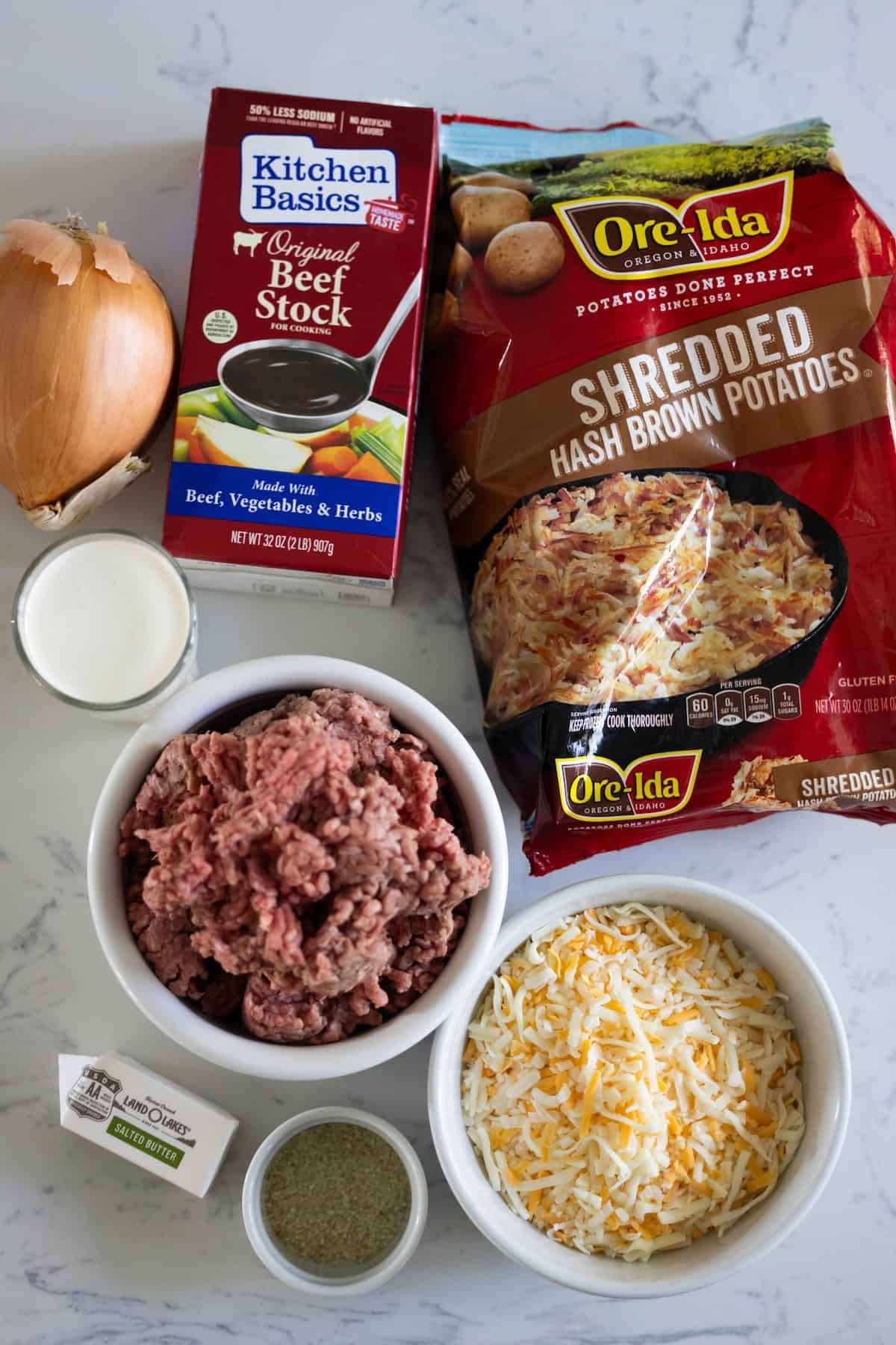 Ingredients on a countertop for a creamy hash brown hamburger soup include onion, beef stock, shredded hash brown potatoes, ground beef, shredded cheese, stick of butter, and a small cup of cream.