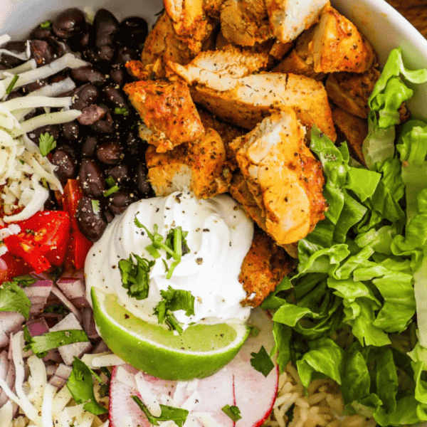 A bowl containing grilled chicken pieces, black beans, shredded cheese, chopped tomatoes, sour cream, lettuce, lime, radish slices, and diced onions, garnished with cilantro—perfect for hearty chicken burrito bowls.
