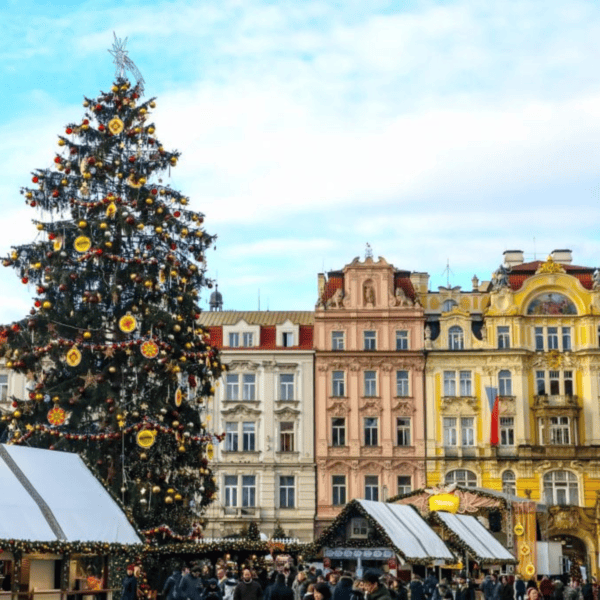 A large Christmas tree decorated with ornaments stands in a bustling Christmas Market, with colorful historic buildings in the background and people gathered around white booths.